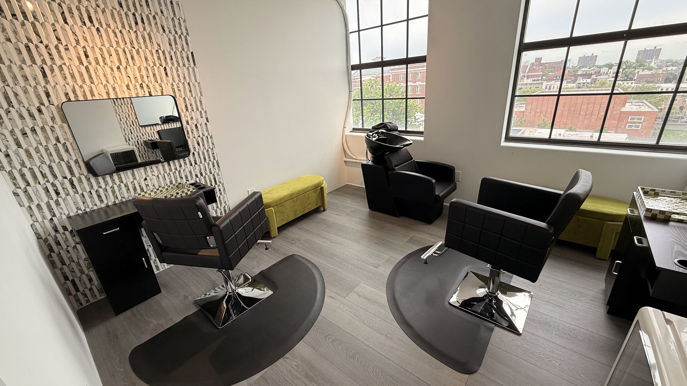 Hair studio: two styling stations with chrome chairs, two black wash basins, mosaic feature wall, yellow velvet bench, and Brooklyn skyline through arched windows